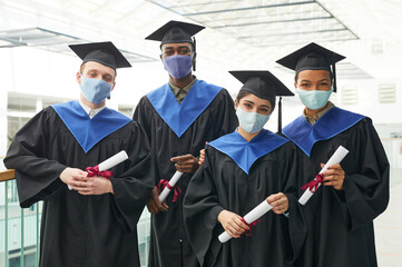 Diverse group of young people wearing graduation gowns and masks while posing indoors in college...