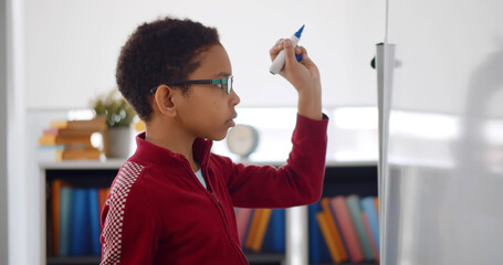 Portrait of african boy writing solution of sums on white board at school.