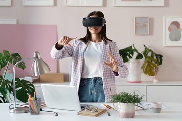 Confident young woman in virtual reality headset pointing in the air while standing at her working place in office