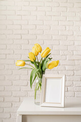 yellow tulips in a glass vase and blank photo frame on a white brick wall background
