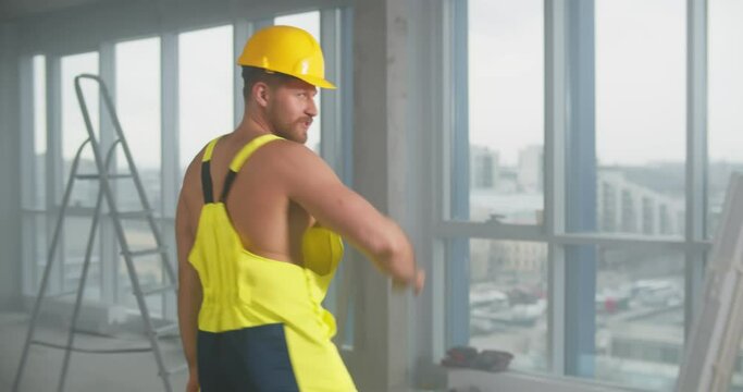 Sexy young shirtless builder in overall and hardhat dancing at construction site