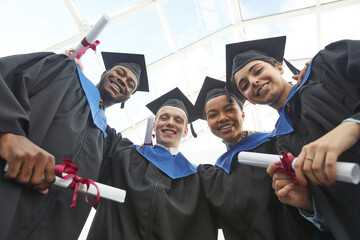 Low angle view at diverse group of happy young people wearing graduation gowns and holding diplomas...