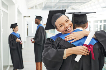 Portrait of happy African-American girl wearing graduation gown hugging friend and smiling at...