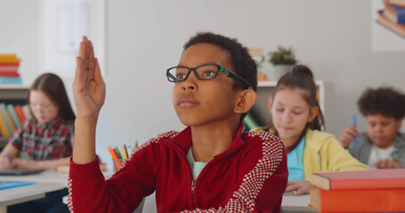 Cheerful african boy raising hand and asking question in classroom.