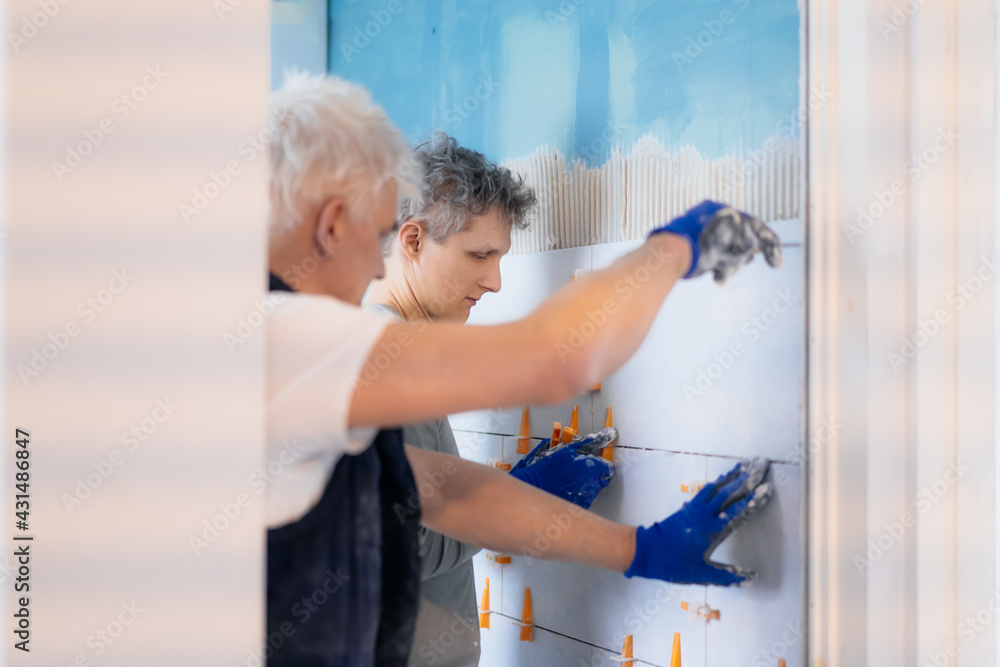 Wall mural two tilers installing wall tile at home. father and son laying tiles on a bathroom wall. diy, do it 