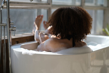 Obraz na płótnie Canvas back view curly black girl laying, relaxing in the bath indoors. young woman relaxing in warm bathtub with foam and bubbles. Tired female enjoying rest pamper herself.
