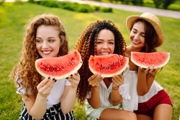 Three young having fun eating watermelon In the park. Young friends laughing and enjoying holidays together. Friendship, youth and travel concept.