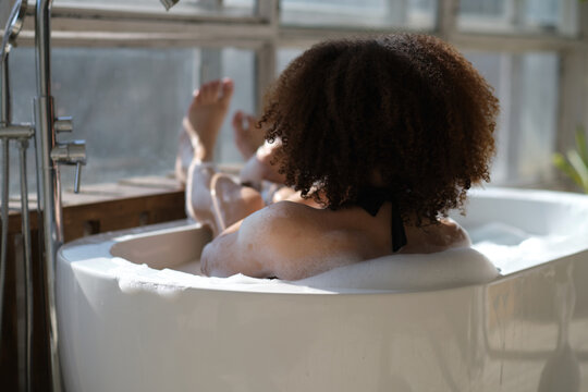 back view curly black girl laying, relaxing in the bath indoors. young woman relaxing in warm bathtub with foam and bubbles. Tired female enjoying rest pamper herself.