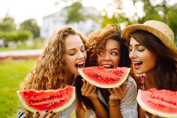 Three young having fun eating watermelon In the park. Young friends laughing and enjoying holidays together. Friendship, youth and travel concept. - obrazy, fototapety, plakaty