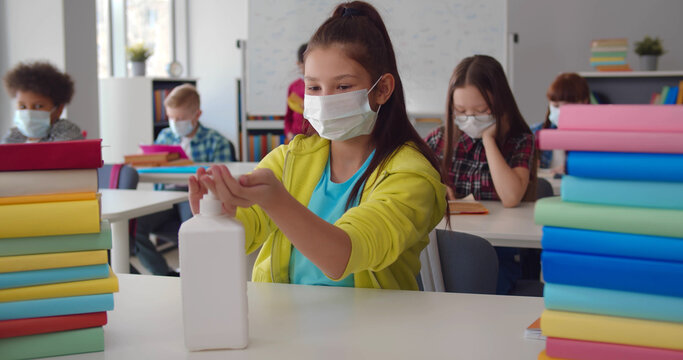 Schoolgirl in protective mask using antibacterial hand sanitizer in classroom