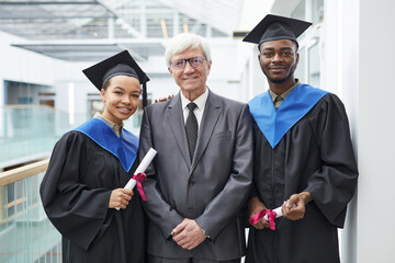 Waist up portrait of two college graduates holding diplomas while posing with mature professor and smiling at camera