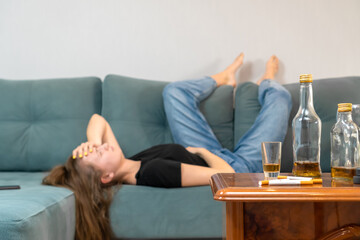 Drunk young dark-haired woman is lying on the couch after a party and holding her head, empty bottles are on the table. Focus on bottles