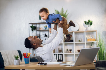 Cheerful african man playing with cute baby boy and using modern laptop for remote work. Young...