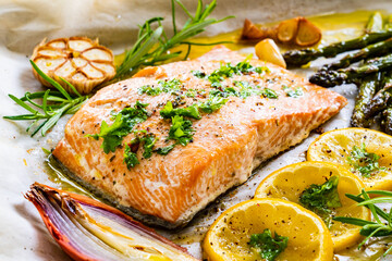 Sheet pan dinner - roasted salmon steak with asparagus, lemon ,rosemary, tomatoes, onion and garlic on cooking pan on wooden table
