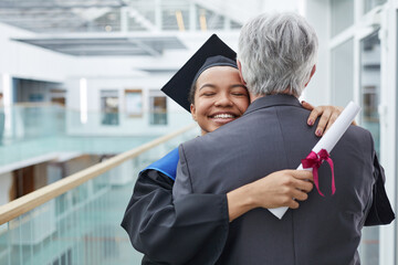 Portrait of happy African-American woman wearing graduation gown hugging father or college...