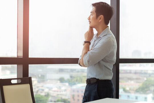 Thoughtful Asian Business Man Standing Next To Big Window Looking Out.