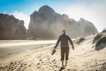 Man walking with his shoes in his hand on the beautiful and mountainous Big Sur Coast of California on a misty yet sunny day, United States of America aka USA.
