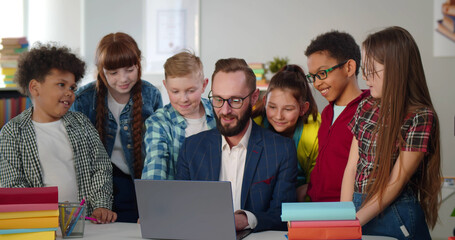 Male teacher in glasses showing scientific video to joyful children using laptop
