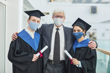 Waist up portrait of two college graduates wearing masks while posing with mature professor indoors