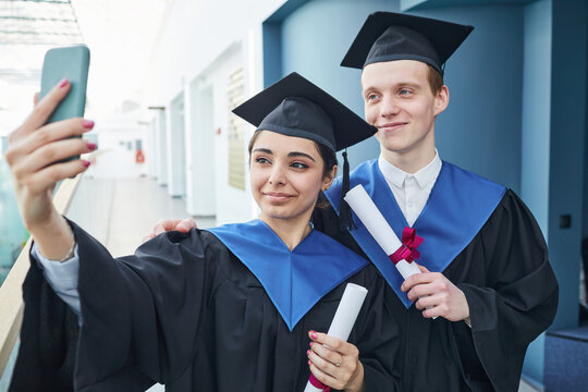 Waist up portrait of two college graduates taking selfie during ceremony indoors, copy space