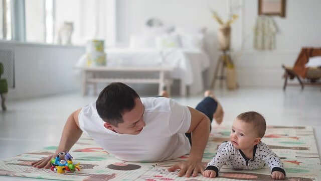 Happy young dad lying on floor carpet, lifting in air adorable little baby son, playing airplane or imagining travel trip together in living room, happy father's day concept. High quality 4k footage