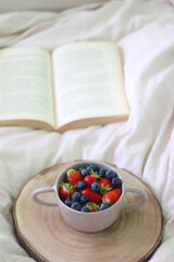 Bowl with blueberries and strawberries and open book on a bed. Selective focus. 