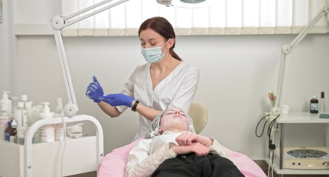 Young caucasian woman in facial mask and gloves doing face treatment to client at modern clinic. Female beautician doctor cosmetologist applying face cleansing powder at beauty parlor. Face wash