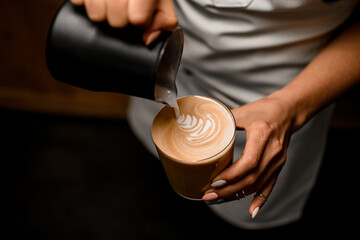 woman barista gently drawing pattern pouring milk into glass with latte drink