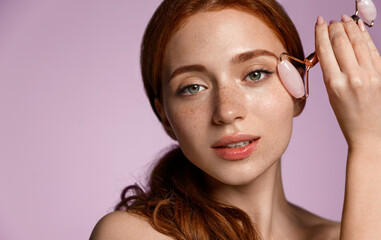 Skincare spa. Close up redhead woman uses natural quarz roller under eye, reduce puffiness and dark...