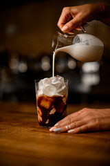 close-up of glass with ice cubes and coffee into which female barista pours milk