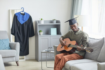 Portrait of young African-American man playing guitar at home while preparing for graduation...