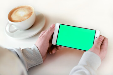 Hand holding green Screen Phone with a Cup of Cappuccino on white table. Woman using Mobile Phone...
