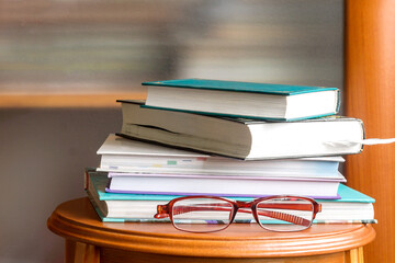 A stack of books on a wooden reading console with female glasses for vision. 