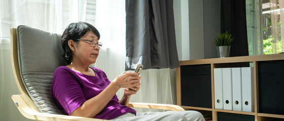 portrait photo of elderly or old Asian retirement woman smiling and using smartphone for social media checking while siting on armchair in living room. technology, communication and people concept
