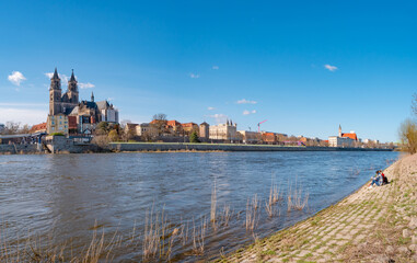Fototapeta na wymiar Panoramic view over Magdeburg historical downtown, Elbe river and the cathedral in early Spring with two girls sitting at the bank with warm illumination and blue sky, Germany.