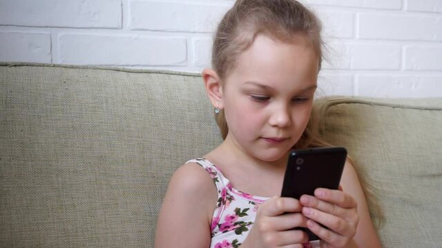 Girl playing mobile game on smartphone on sofa. Preschooler playing mobile phone. Kid using phone for gaming. Child playing video game at home