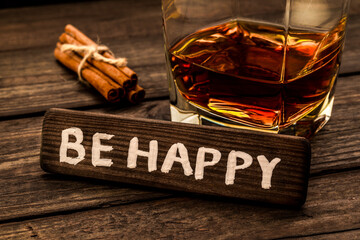 Glass of brandy with cinnamon sticks tied with jute rope and the wooden plank on it is an inscription "BE HAPPY" on an old wooden table. Close up view, focus on the inscription