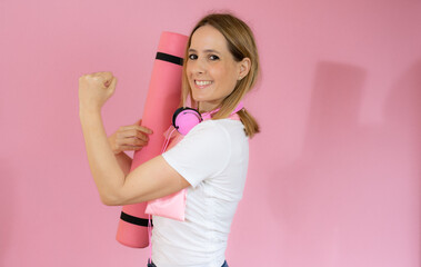 young woman holding a pilates mat and wearing headphone with a smile - isolated on pink