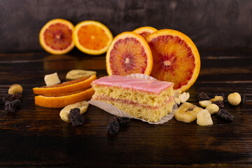 sponge cake in pink glaze with slices of red orange and a scattering of dried fruits and nuts on a wooden background close-up