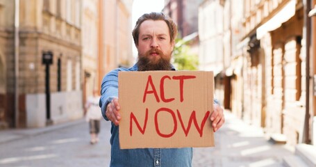 Portrait of Caucasian young man standing outdoors in town and showing table Act Now. Male demonstrating board with protest against pandemic, political or environmental issues. Single protest.
