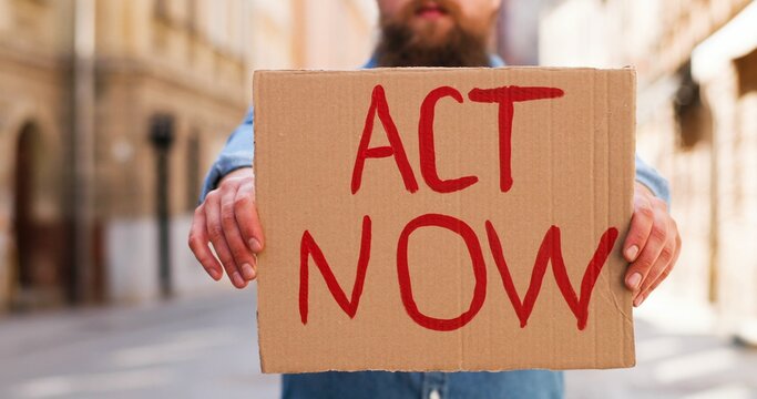 Close Up Of Caucasian Young Man Standing Outdoor In Town And Showing Table Act Now. Male Demonstrating Board With Protest Against Pandemic, Political Or Environmental Issues.