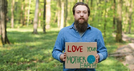 Portrait shot of Caucasian young male eco acivist standing in green forest and holding poster with...