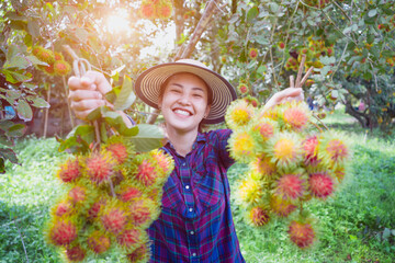A lovely young woman wearing a plaid shirt and a standing hat, holding a bright red rambutan and smiling happily, the colorful rambutan is ripe and ready to eat in a natural garden in Thailand.