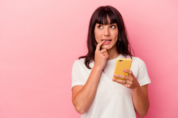 Young caucasian woman holding a mobile phone isolated on pink background relaxed thinking about something looking at a copy space.