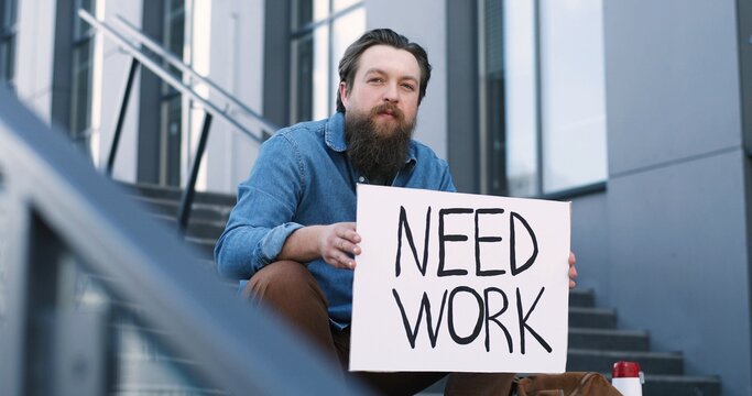 Portrait of young workless Caucasian man with beard sitting on stairs outdoors and holding carton table Need Work. Unemployment after pandemic lockdown. Workless male.