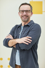 Vertical portrait of mature male teacher smiling at camera while standing with arms crossed in school classroom © Seventyfour