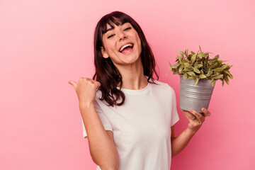Young caucasian woman holding a plant isolated on pink background points with thumb finger away,...