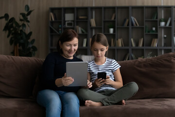 Modern elderly grandmother and teenage granddaughter relax at home use gadgets together. Happy...