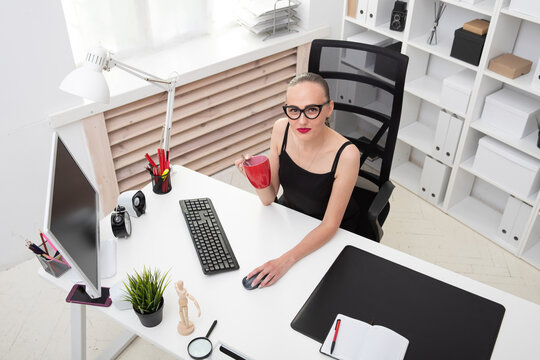 Portrait of a beautiful girl in a black blouse who drinks coffee from a red mug at the workplace