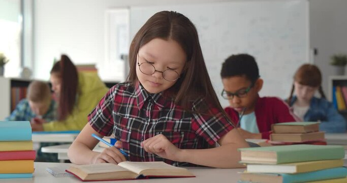 Smart schoolgirl sitting in classroom and writing in textbook